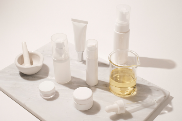 Skincare routine products on white counter with grey marble cutting board: bottles, mortar and pestle, and golden serum.